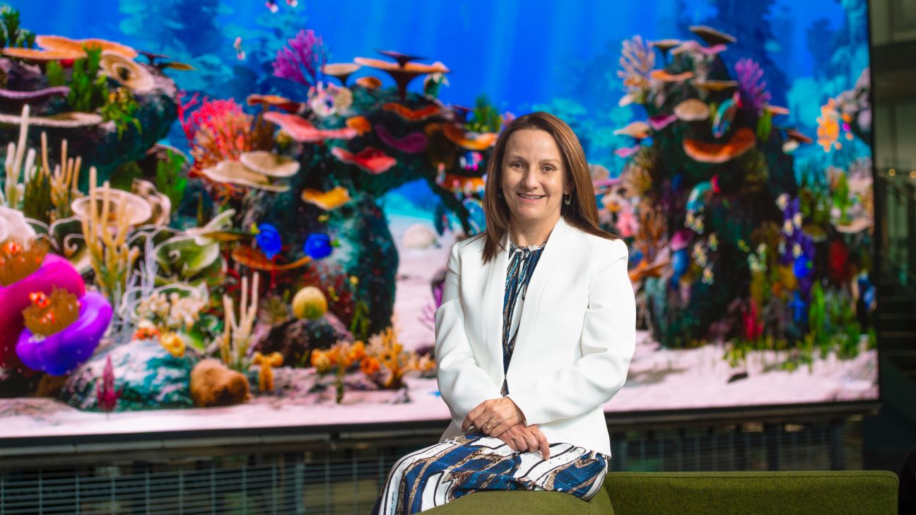 A woman with brown hair in a white blazer and blue-and-white dress sits in front of a colourful aquarium backdrop.