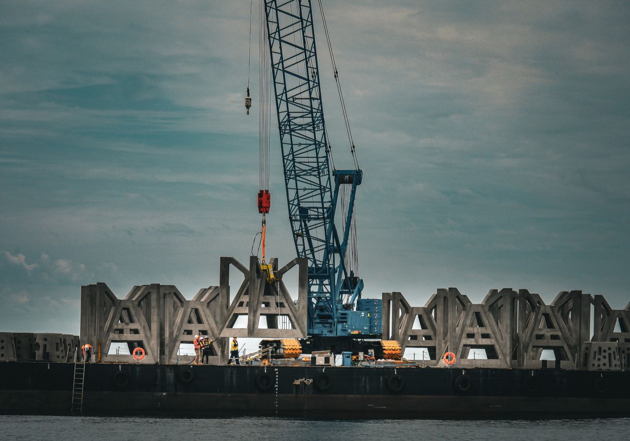 Large crane lifting precast concrete structures on a barge, with several workers in high-visibility gear standing along the deck above calm water under a cloudy sky. 
