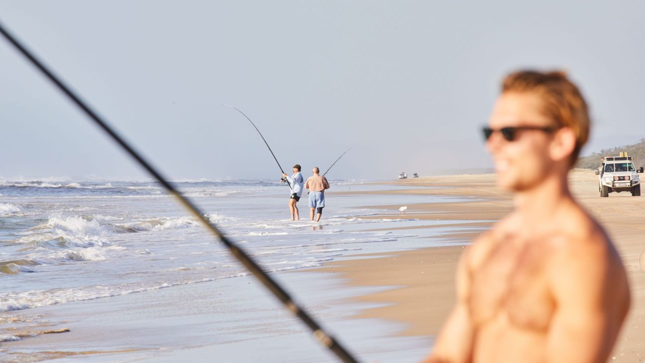 People fish from the shoreline of a long sandy beach beside rolling ocean waves. In the foreground, a fishing rod and a person wearing sunglasses are out of focus, while two anglers stand in the shallow surf with rods cast into the water. A four-wheel-drive vehicle is parked further along the beach under clear skies.