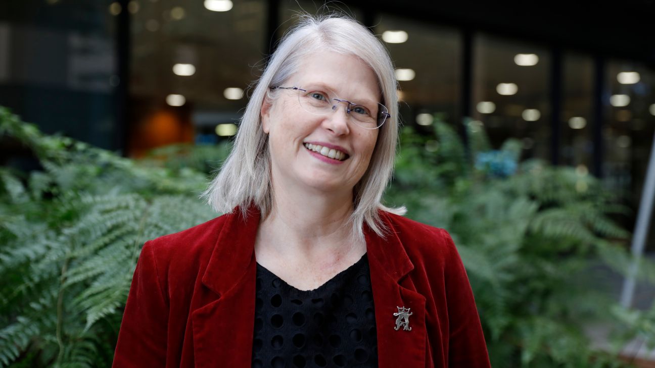 A woman with white-blonde shoulder length hair and clear framed glasses, wearing a black top and a red velvet blazer.