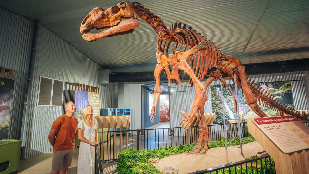 Two adults gaze up at a large dinosaur skeleton replica enclosed by a small fence.