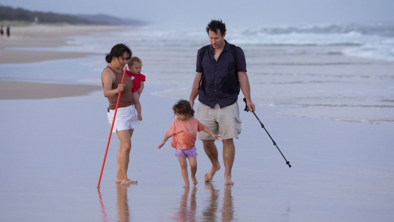 Three people stand barefoot on wet sand at the edge of a beach, with ocean waves rolling in behind them. One adult holds an infant and a brightly coloured dingo safety stick, while another adult holds a walking pole. A young child stands between them near the waterline. The reflective sand, overcast sky and long stretch of beach create a calm coastal scene.
