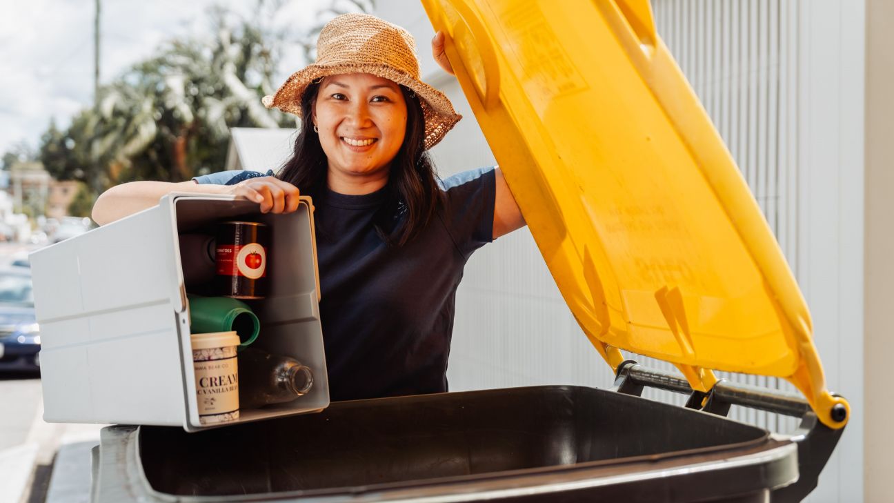 A person standing beside a wheelie bin lifts the yellow lid while emptying a grey kitchen recycling caddy containing cans, plastic containers and bottles into the bin. The scene is outdoors in daylight, with trees and parked vehicles visible in the background.