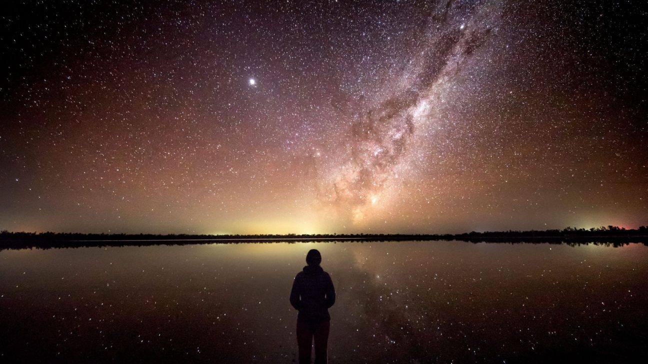 A person stands at the edge of a still lake beneath a dazzling night sky filled with countless stars and the bright band of the Milky Way. The calm water reflects the stars and galaxy almost perfectly, creating a mirror-like effect that stretches across the landscape and highlights the area's dark sky conditions.
