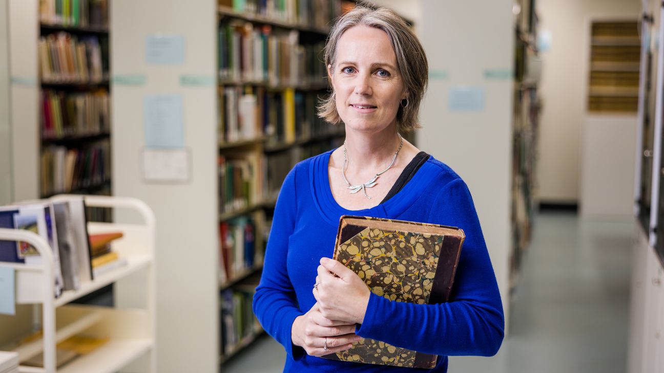 A woman with short grey hair wearing a blue sweater holds a brown paisley pattered notebook in her arms.