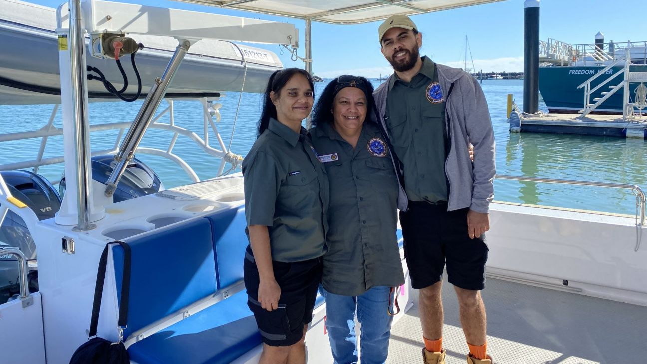 Three people smiling on a boat: two women and a bearded man in uniform shirts, posing together on a sunny day with water and boats in the background.