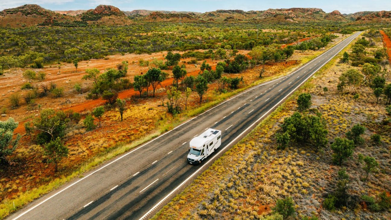 A large white caravan drives down a long road in the middle of a sparse, outback scene.