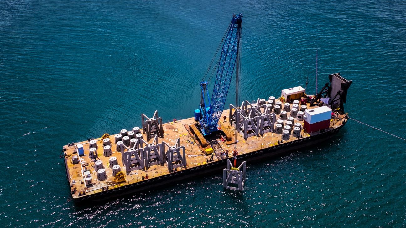 Aerial view of a barge on open water carrying a large blue crane and numerous precast concrete artificial reef modules, with a structure being lifted over the side into the sea. 