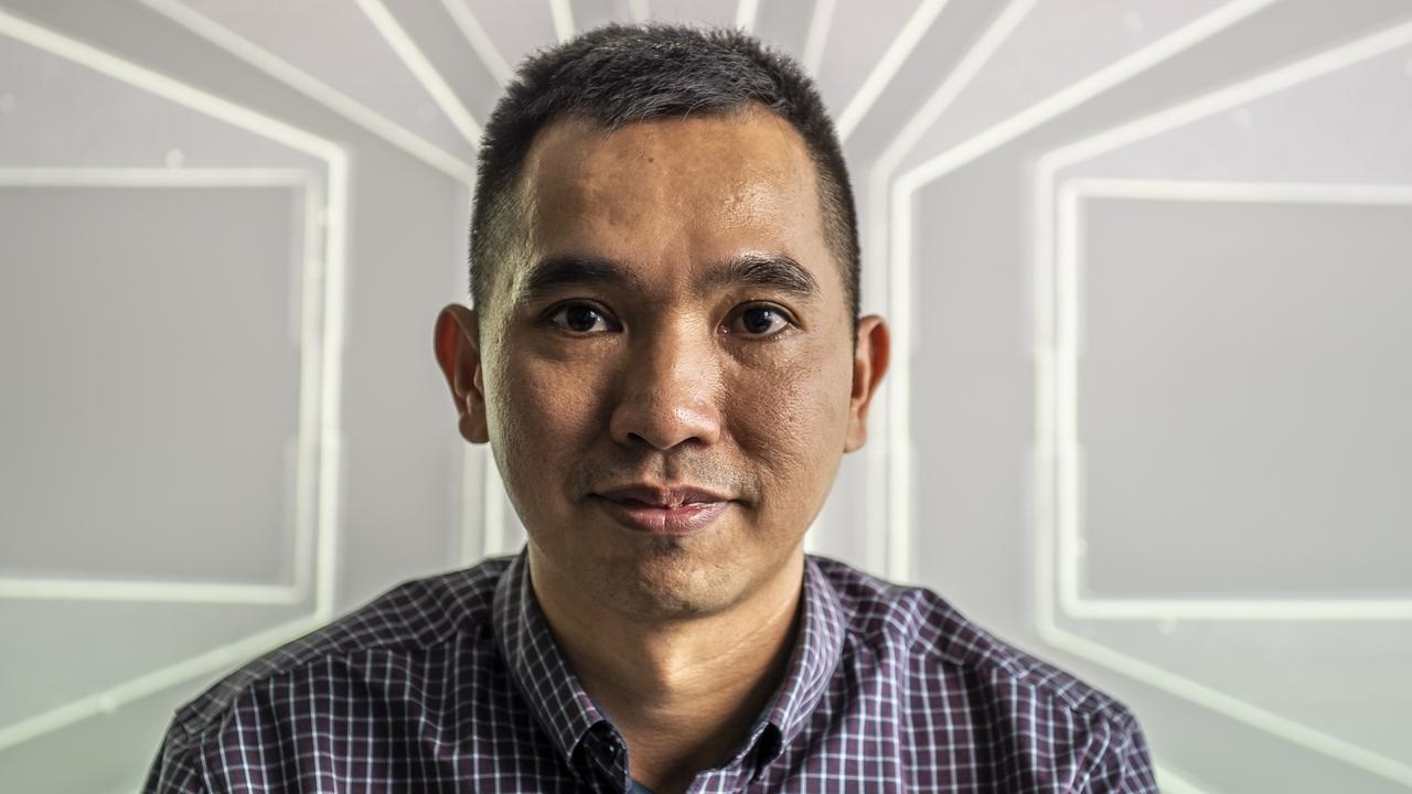 Headshot of a man with short dark hair wearing a patterned button-up shirt in front of an abstract background.