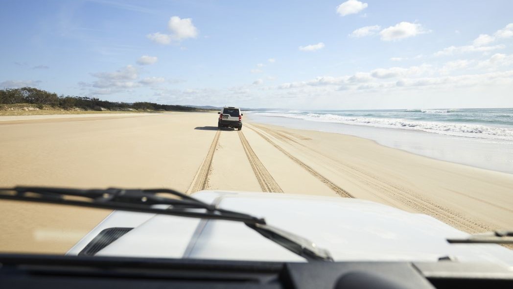 View from inside a four-wheel-drive vehicle travelling along a wide sandy beach. Another four-wheel-drive is driving ahead, leaving tyre tracks in the firm sand beside the shoreline. Ocean waves roll in on the right, while low dunes and coastal vegetation line the beach under a bright sky with scattered clouds.
