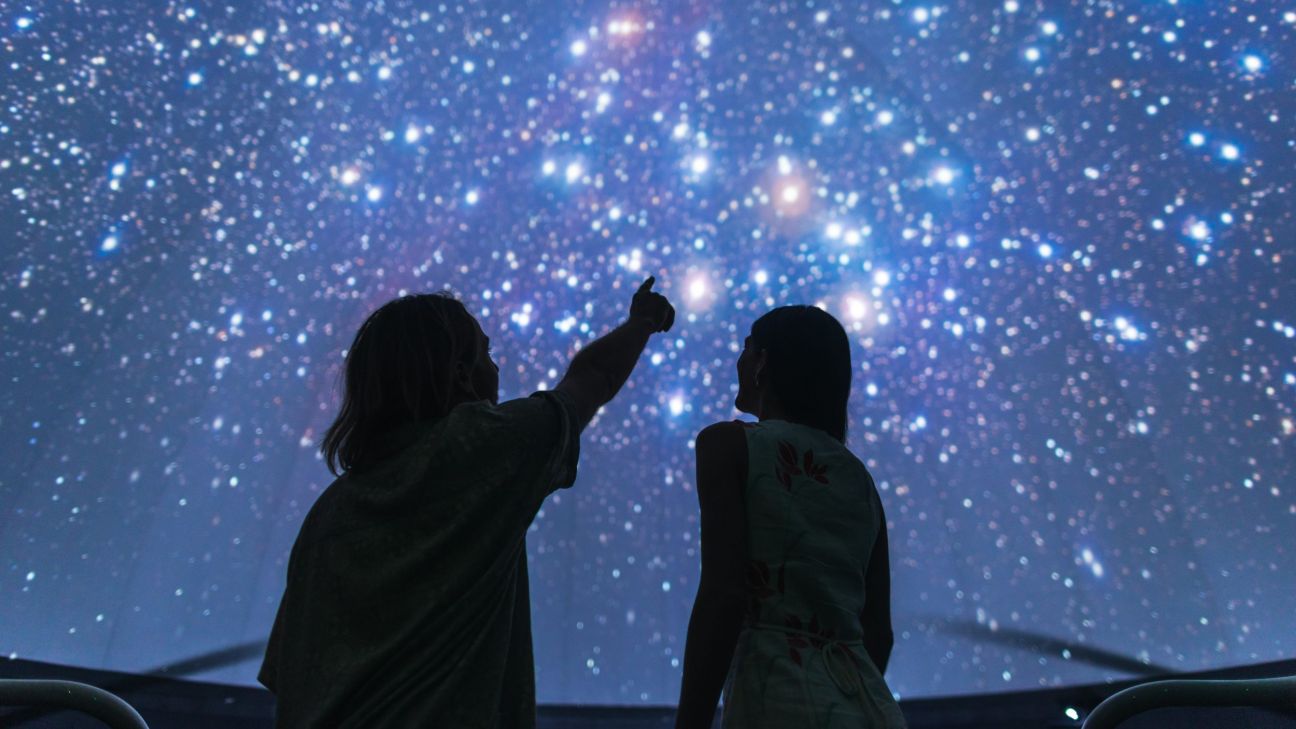 Two people sit beneath a large domed projection of a star-filled night sky, with one person pointing toward a cluster of bright stars. The immersive display shows thousands of stars and galaxies across the curved ceiling, creating the experience of exploring the night sky inside a planetarium or astronomy centre.