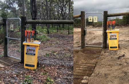 A yellow bin with several orange-tipped sticks poking out from the top. The bin is next to a tall fence with a chain-link gate surrounded by brown leaves and bark and another yellow bin with several orange-tipped sticks poking out from the top. The bin is next to a tall fence with a chain-link gate on a sandy beach with a wooden boardwalk-style path.