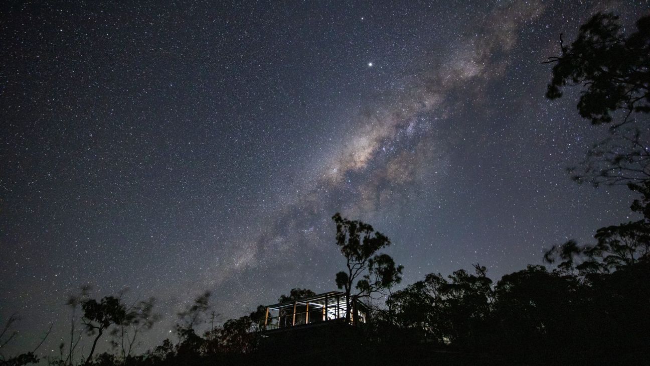 A viewing platform surrounded by trees sits beneath a remarkably clear night sky, illuminated by soft light from within. The Milky Way stretches diagonally across the sky in a dense band of stars, towering above the forested landscape and showcasing the remote area's exceptional stargazing conditions.
