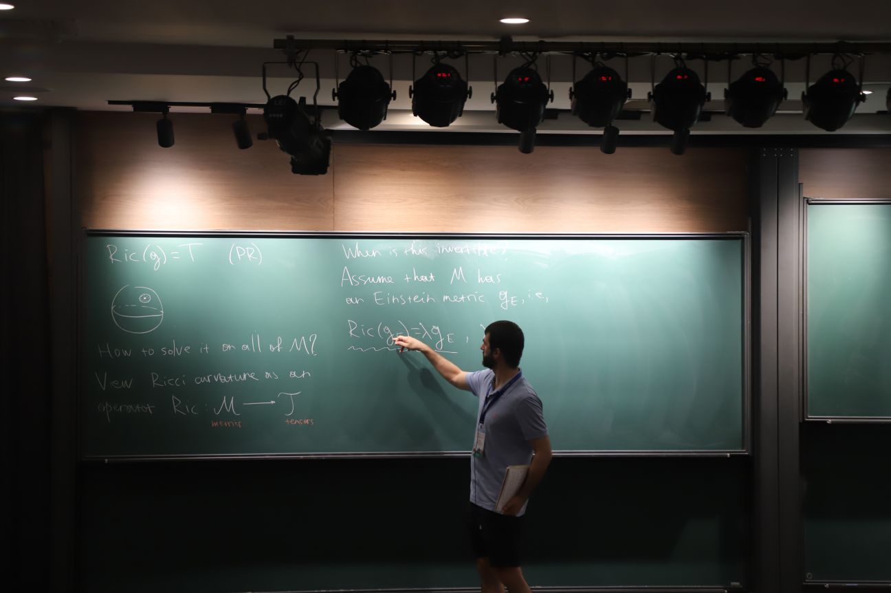A man in a blue polo shirt pointing at equations on a blackboard with spotlights beaming down.