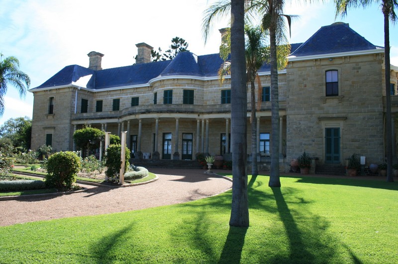A large, grand, sandstone two-storey mansion sits towards the back of a green lawn and driveway.