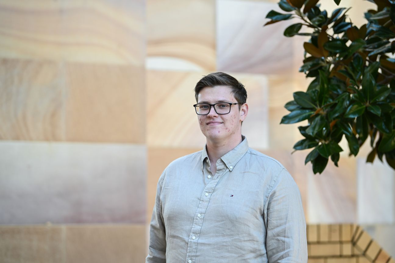 A headshot of a young man with glasses wearing a beige long-sleeved collared shirt standing against a sandstone wall background.