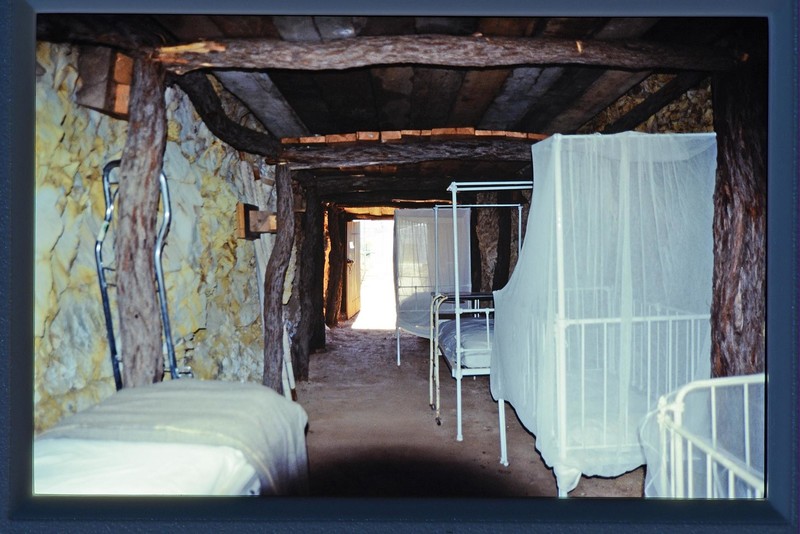 Old, white single beds rest against the follow of an underground cave with stone walls and a wooden roof.
