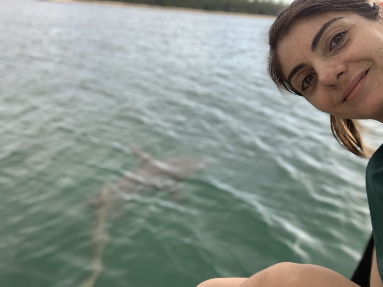 A selfie-style image of a woman to the right of the frame with water in the back – she is using her hand to highlight a shark in the water behind her.