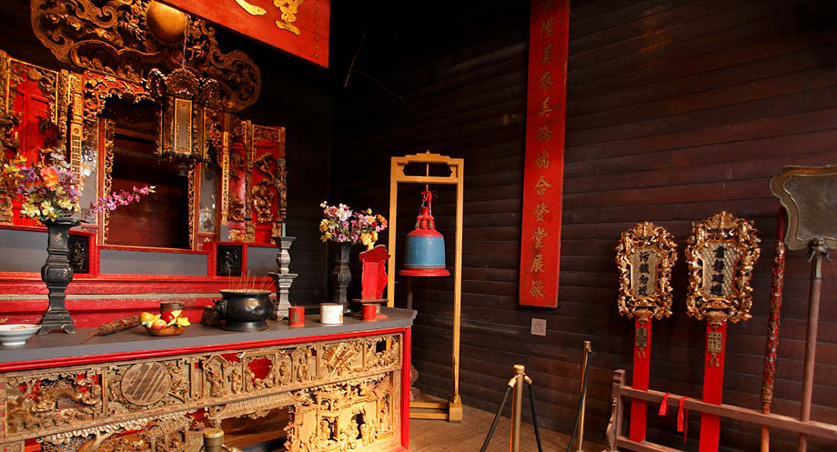 View inside the Hou Wang Chinese Temple - an ornate carved counter, blue and red bell, red banners, and flowers in black vases are visible.