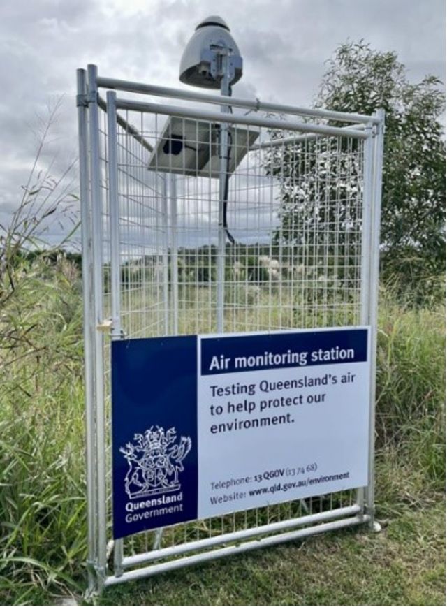 A fenced-off air monitoring station in a grassy area, with a sign from the Queensland Government stating, 