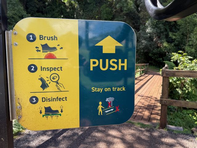 Instructional sign at a nature trail entrance showing steps: Brush, Inspect, and Disinfect shoes; push gate to enter; reminder to stay on track. A wooden bridge and greenery are in the background.