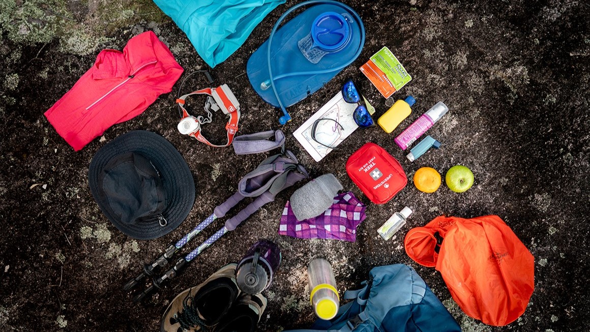 A flat lay of essential hiking gear displayed on the ground, including a hydration pack, rain jackets, a first aid kit, a map, trekking poles, a hat, sunglasses, snacks, and other safety essentials.