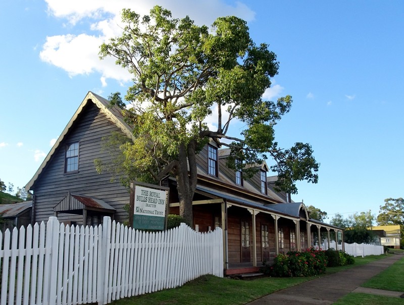 An old, sprawling wooden inn with a tall white picket fence and a large tree out the front.