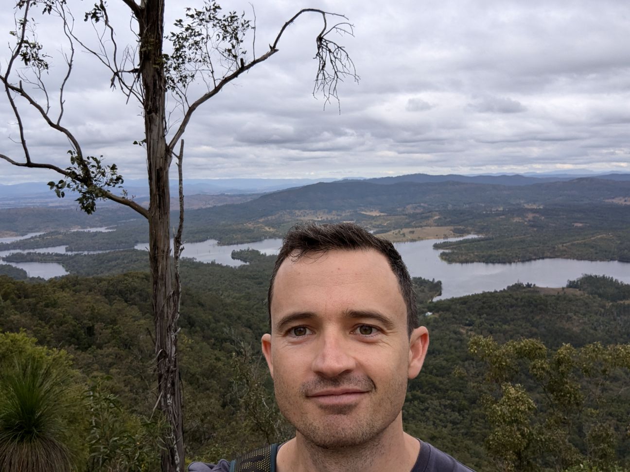 A selfie-style image of a man with short brown hair with a river, greenery, and trees in the background.