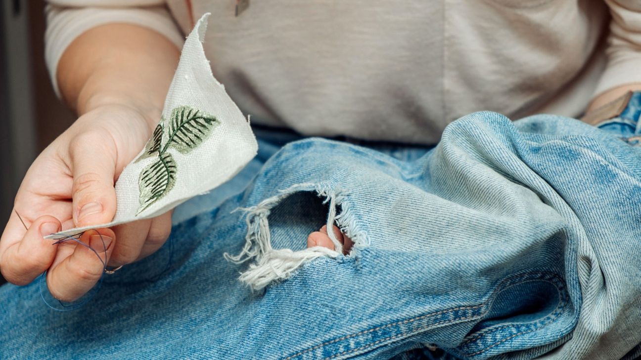 Close-up of a person holding a pair of jeans with a hole in them in their lap. They have a fabric patch for mending in their other hand.