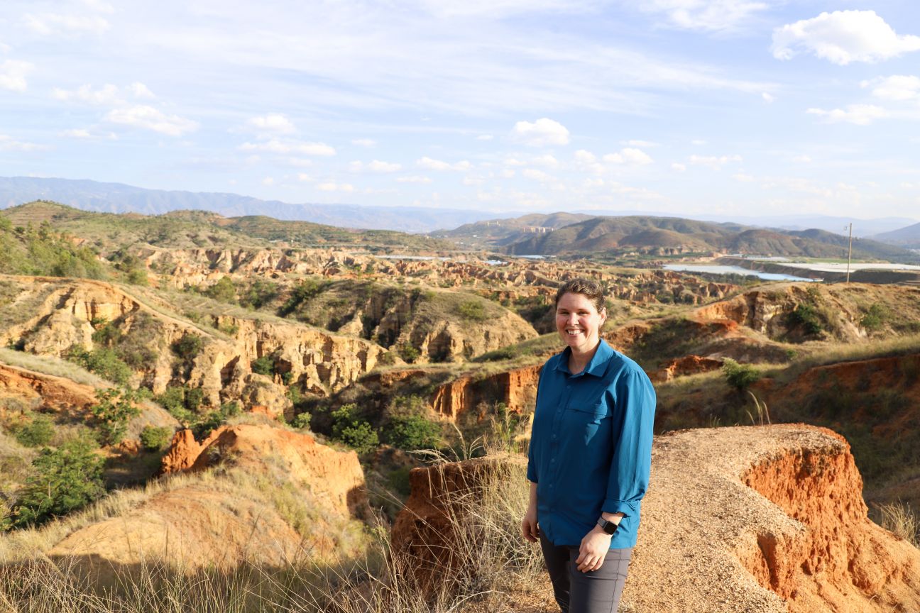 A woman in a blue long-sleeved shirt and grey pants standing amongst green and brown rocky formations.
