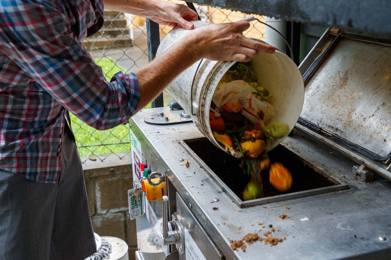 A man in a checked shirt empties a bucket of colourful food scraps into a large metal composting receptable.