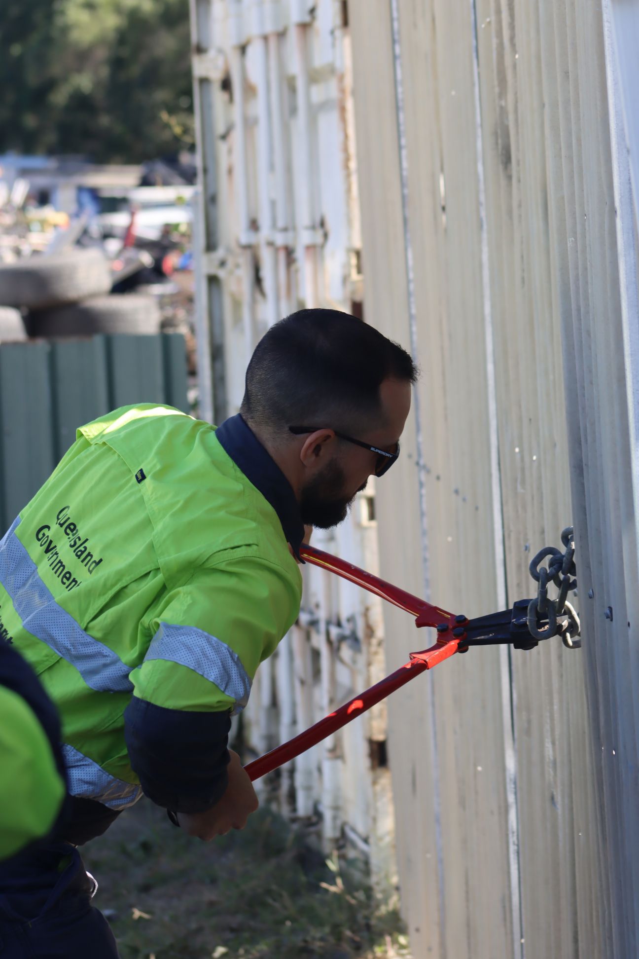 A man in hi-viz Queensland government uniform using bolt cutters to cut a chain in a row of shipping containers