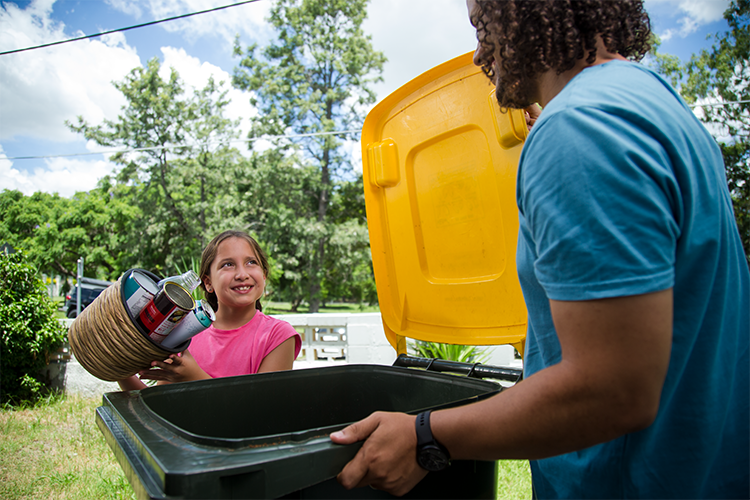 A young girl in a pink shirt tips a container full of loose recycling into a yellow lid bin.