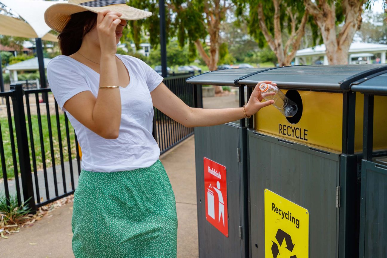 A woman in a white t-shirt, green skirt, and hat puts a clear plastic bottle into a public recycling bin.